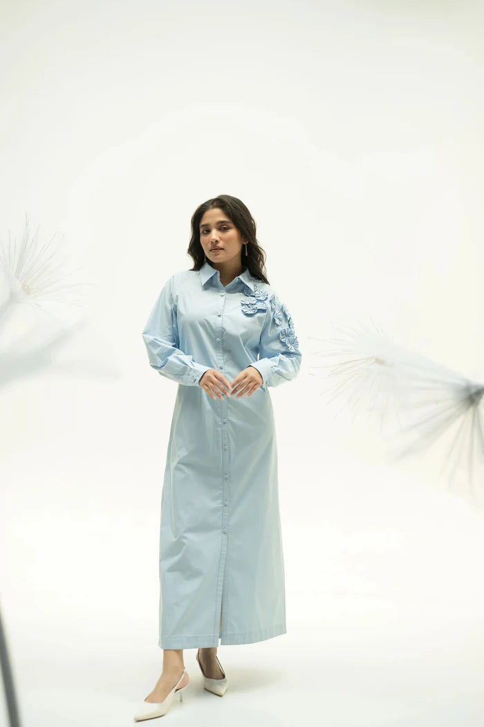Woman wearing light blue midi dress with corsage detail standing in studio with soft white background.
