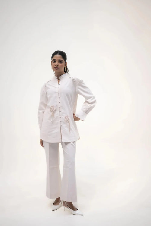 A model standing with a hand on her hip, wearing a long white shirt and white trousers, in a neutral pose against a soft background.