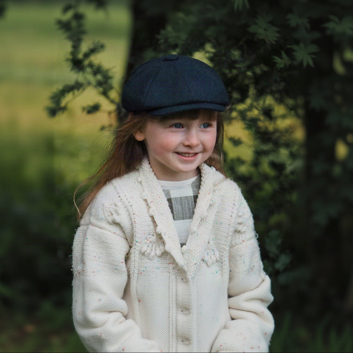 A young child with long hair smiling, wearing a blue tweed newsboy cap and a white knit cardigan.