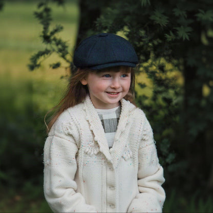 A young child with long hair smiling, wearing a blue tweed newsboy cap and a white knit cardigan.