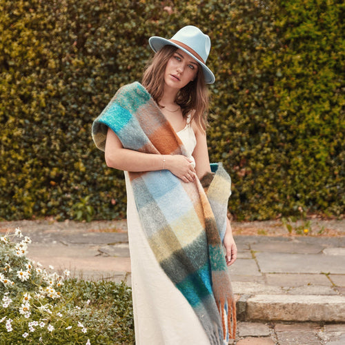 A woman wearing a sky blue felt fedora hat and a color block blanket scarf with blue and brown hues.