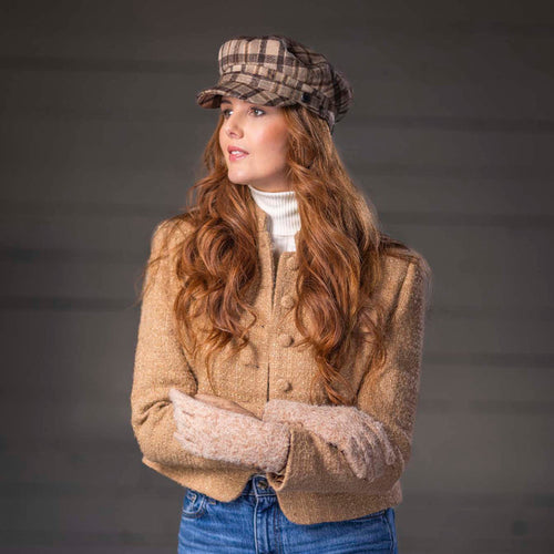 A woman wearing a beige cropped knit cardigan, blue jeans, a white turtleneck, and a brown plaid newsboy cap, standing against a dark gray background.